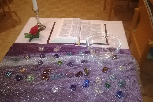 Pöydällä violetti liina, jonka päällä erivärisiä lasikiviä. Raamattu, kynttilä ja ruusu valkoisella alustalla.